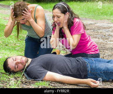 Young man lying down with medical emergency, two young women acting hysterically, outdoors environment Stock Photo