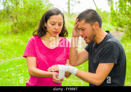 Young man with injured wrist sitting and getting bandage compression wrap from female, outdoors environment Stock Photo