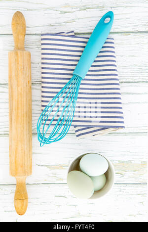 Eggs in a ceramic bowl with a whisk and rolling pin on a rustic white painted wood table top shot from above Stock Photo