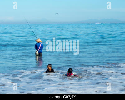 A man standing in the sea fishing while two young boys are playing closer to shore. Stock Photo