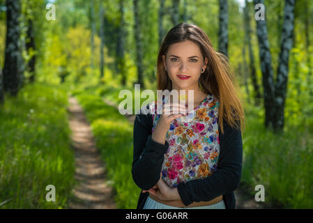 Young woman with red lips and brown hair is holding one yellow dandelion in hand while standing on a footpath in a forest during Stock Photo