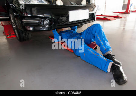 Mechanic in blue uniform lying down and working under car at the garage. Stock Photo