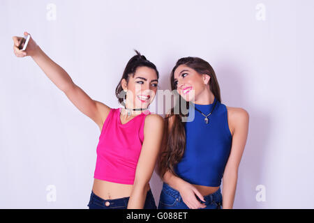 Two stylish women taking a selfie with a mobile phone. Best friends having fun, in urban outfit, isolated over grey background. Stock Photo