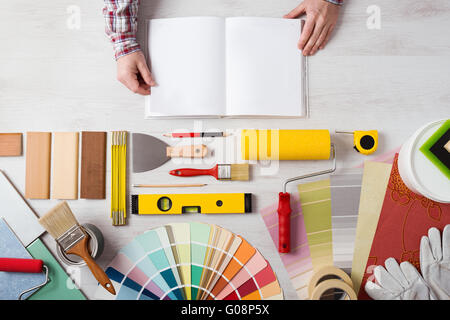 Hands holding DIY training open manual with work tools, color swatches and painting rollers at bottom, top view Stock Photo