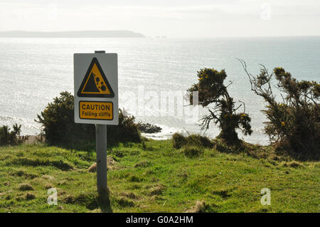 Caution -falling cliffs - warning sign on pathway above a sunlit sea - bushes  on the edge - branches like desperate waving arms Stock Photo