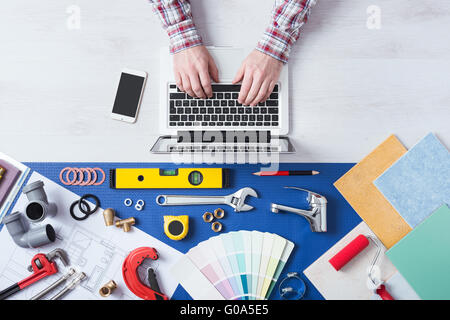 Male hands using a laptop next to plumbing work tools, tiles and swatches, online booking and home plumber service Stock Photo