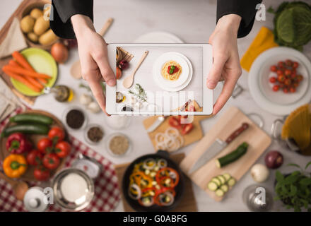 Cook's hands holding a touch screen tablet close up, kitchen table with food ingredients, vegetables and utensils on background, Stock Photo