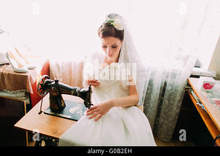 Beautiful young bride makes last minute adjustments to her Wedding Dress at an ancient old treadle sewing machine Stock Photo