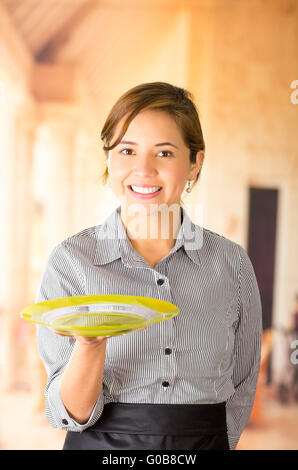 Young brunette waitress wearing uniform with friendly smile, holding up tray using one hand, posing happily for camera Stock Photo