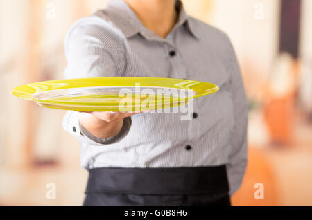 Closeup young brunette waitress wearing uniform, holding up yellow empty tray using one hand Stock Photo