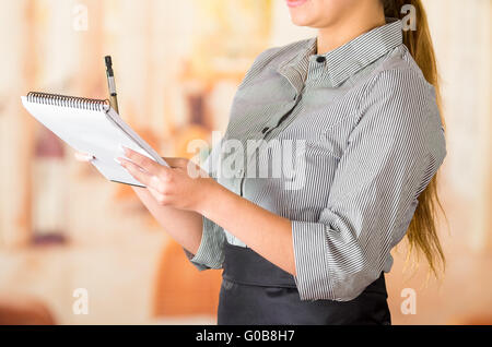 Young brunette waitress wearing uniform taking order writing on notepad, restaurant background Stock Photo