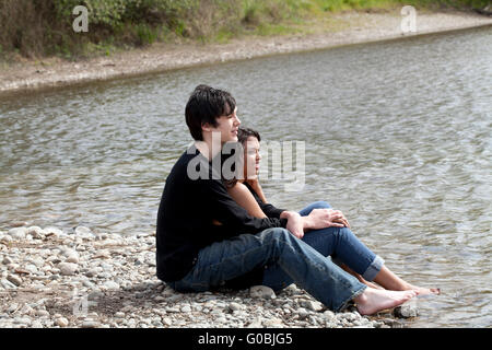 teen boy and girl sitting holding each other on river bank Stock Photo