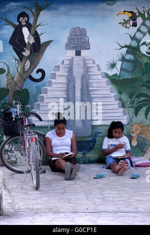 Local schoolgirls sit and  study in front of a painted wall depicting Mayan ruins of Tikal in the village of El Remate at the shore of Lake Peten Itza in the northern Peten Department in Guatemala. Central America Stock Photo