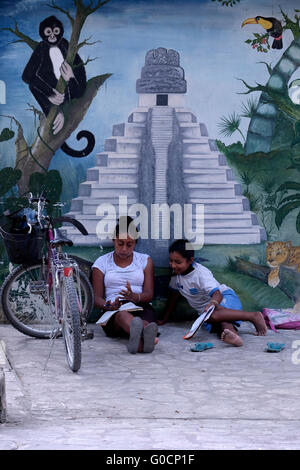 Local schoolgirls sit and  study in front of a painted wall depicting Mayan ruins of Tikal in the village of El Remate at the shore of Lake Peten Itza in the northern Peten Department in Guatemala. Central America Stock Photo