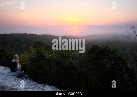 A solo female hiker watching sunrise over the tropical jungle from the ancient Mayan temple number 4 at the archaeological site of Tikal an ancient urban center of the pre-Columbian Maya civilization located in the archaeological region of the Peten Basin in northern Guatemala Stock Photo