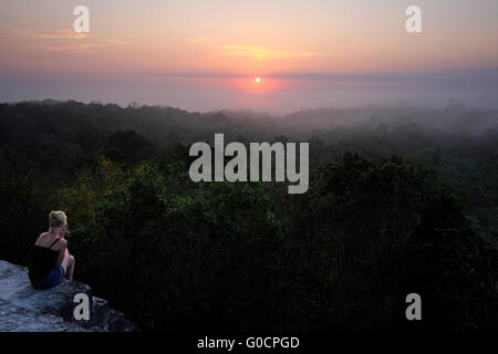 A solo female hiker watching sunrise over the tropical jungle from the ancient Mayan temple number 4 at the archaeological site of Tikal an ancient urban center of the pre-Columbian Maya civilization located in the archaeological region of the Peten Basin in northern Guatemala Stock Photo