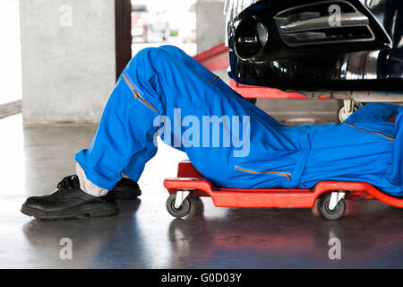 Mechanic in blue uniform lying down and working under car at auto service garage Stock Photo