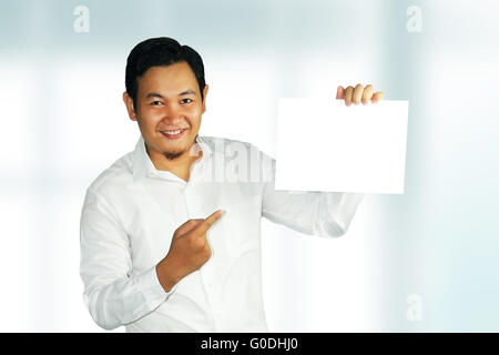 Image of a young Asian male smiling and holding white empty paper over bright background Stock Photo