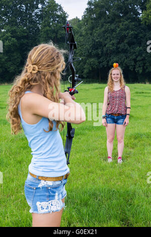 Girl aiming arrow of compound bow at apple on head Stock Photo