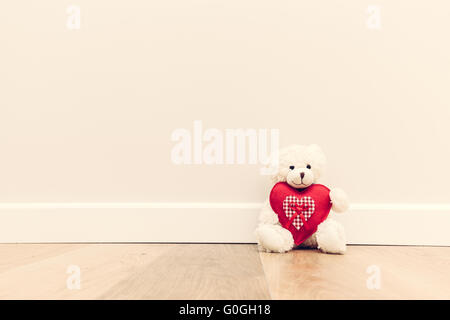 Cute teddy bear with big red plush heart. Sitting on wooden floor against white wall. Stock Photo