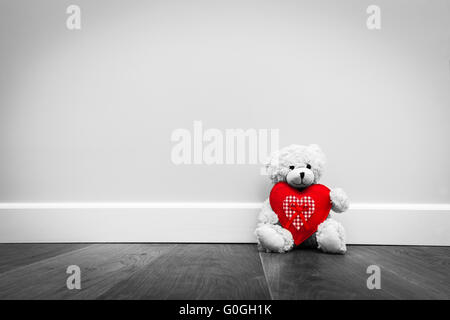 Cute teddy bear with big red plush heart. Sitting on wooden floor against white wall. Stock Photo