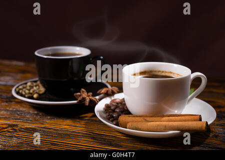 Side view on steaming hot black and white cups of coffee with beans and herbs in saucer on wooden table Stock Photo