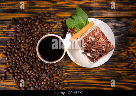 Top down view on delicious square shaped cake slice, spices and leaves in plate beside full cup of coffee surrounded by dark beans over stained wooden background. Stock Photo