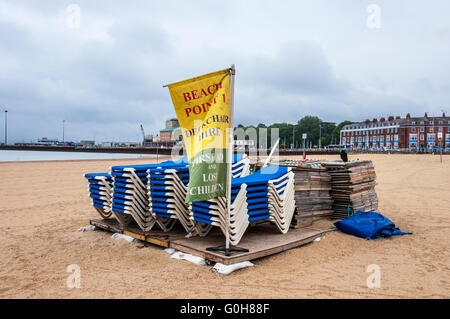 Neatly stacked blue sun loungers and wooden deckchairs rest on duckboards on a nearly deserted Weymouth beach bordered by houses Stock Photo