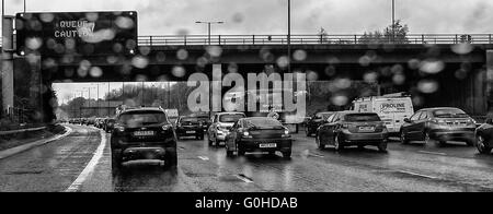 Queuing traffic on motorway with overhead matrix sign stating queue caution slightly blurred by raindrops on windscreen Stock Photo