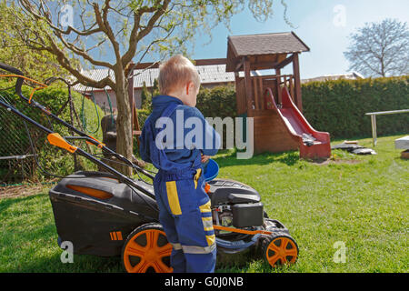 Cute, little boy mows the lawn. Boy inspects the mower, before driving or left engine oil. Stock Photo