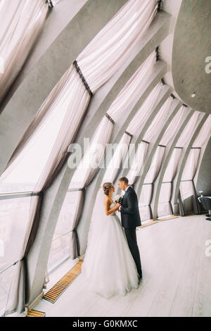 Beautiful married couple facing each other and embracing in a white room with large windows Stock Photo