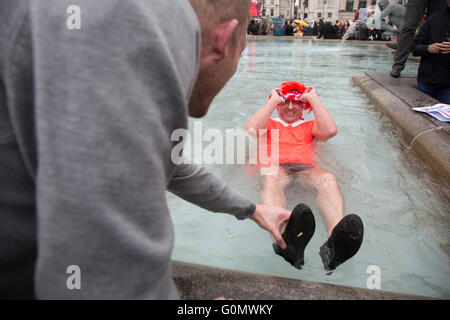 Stag party humiliation pranks as the groom is dressed up as a woman in an orange dress and wig then pushed into the fountains in Trafalgar Square in London, England, United Kingdom. There is a strong tradition at the British Stag Do, to play tricks on the stag at least once. In this case, he had no idea where he was headed, and so blindfolded was perched on the waters edge and thrown in. The stag seemed not to mind but was also, somewhat the worse for wear. Stock Photo