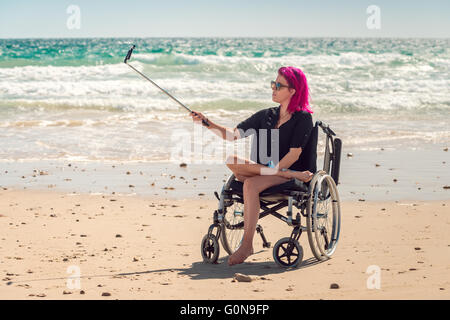 Disabled woman in the wheelchair at the beach taking selfie photos with her mobile phone on selfie stick Stock Photo