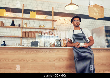 Portrait of young man working at a restaurant wearing an apron and hat leaning to the bar counter. Cafe owner standing at counte Stock Photo