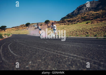 Young man and woman skateboarding with smoke bomb on the road. Young couple practicing skating on a open road. Stock Photo