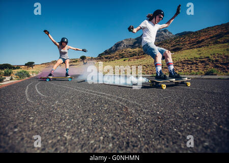 Full length shot of young man and woman outdoors longboarding down the road. Skate boards with smoke grenade. People practicing Stock Photo