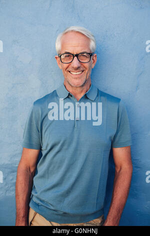 Portrait of handsome mature man with glasses looking at camera and smiling, he is leaning to against a blue wall. Stock Photo