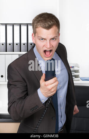 Young businessman is shouting into the phone while standing in the office. A shelf is in the background. The man is looking to t Stock Photo