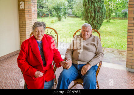 elderly couple holding hands while sitting on wicker chairs on the patio of their home Stock Photo