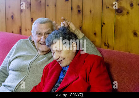 elderly couple kidding while sitting on the sofa in their living room, the man make horns sign on the head of his wife Stock Photo