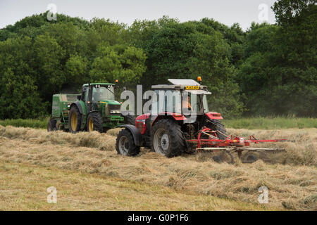 Two farm tractors silage making in a field at Great Ouseburn, North Yorkshire, England - the green one pulling a baler, the red one hauling a rake. Stock Photo