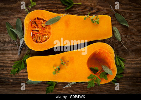 Fresh butternut squash cut in half with sage and parsley on brown wooden background, from above Stock Photo