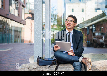 Young smiling asian business man holding a digital tablet sitting outside on city street looking away Stock Photo