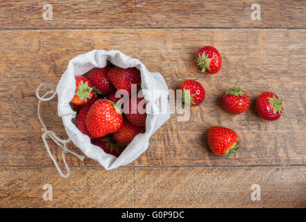 Overhead view of fresh strawberries in a small hessian bag on a wooden kitchen table with a loose strawberries beside it Stock Photo