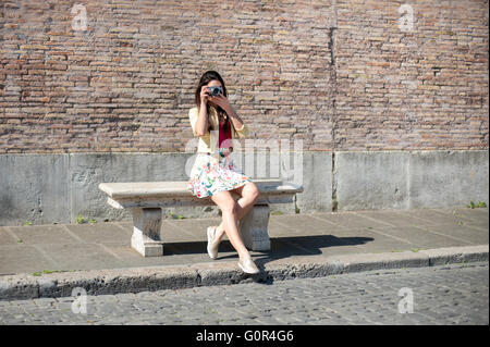 Young tourist woman taking picture outdoor sitting on a bench and wall background Stock Photo