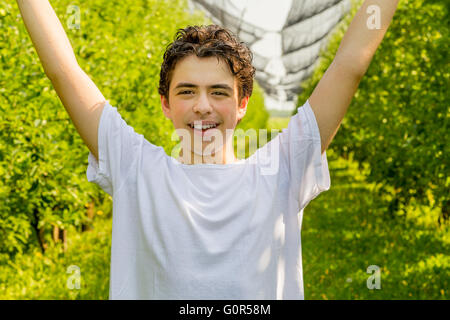 teenager next to rows of apple trees raises his arms under hail net Stock Photo