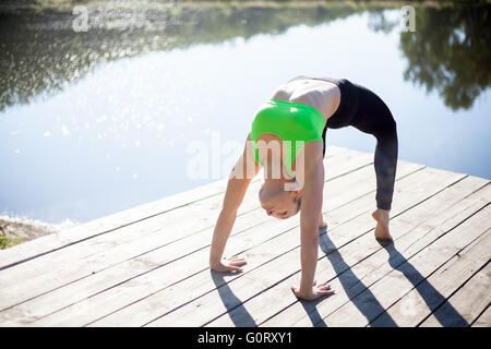 Beautiful sporty fit blond young woman in green sportswear working out outdoors on wooden pontoon on the lake, doing Bridge Pose Stock Photo