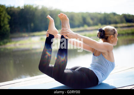 Beautiful sporty fit blond young woman in sportswear working out outdoors in park on lake, doing backbend exercises Stock Photo