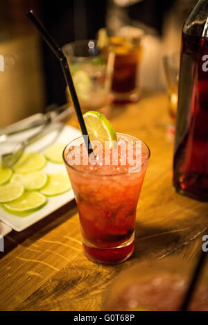 A icy red cocktail with lime and a straw on a wooden bar with slices of lime in the background. Stock Photo