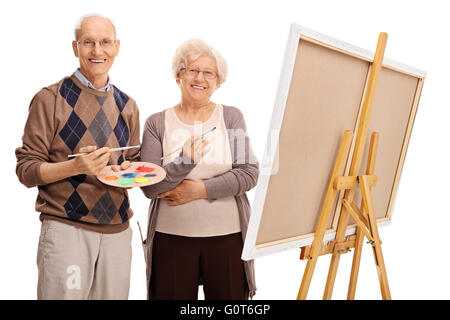 Elderly couple holding paintbrushes and posing next to a canvas isolated on white background Stock Photo